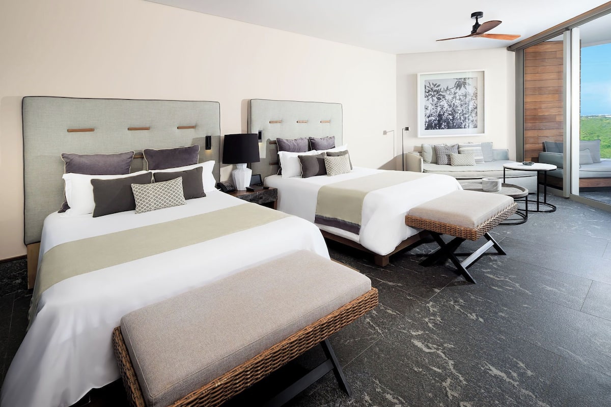 Habitación de hotel con dos camas, un sofá, una mesita y vistas al exterior.