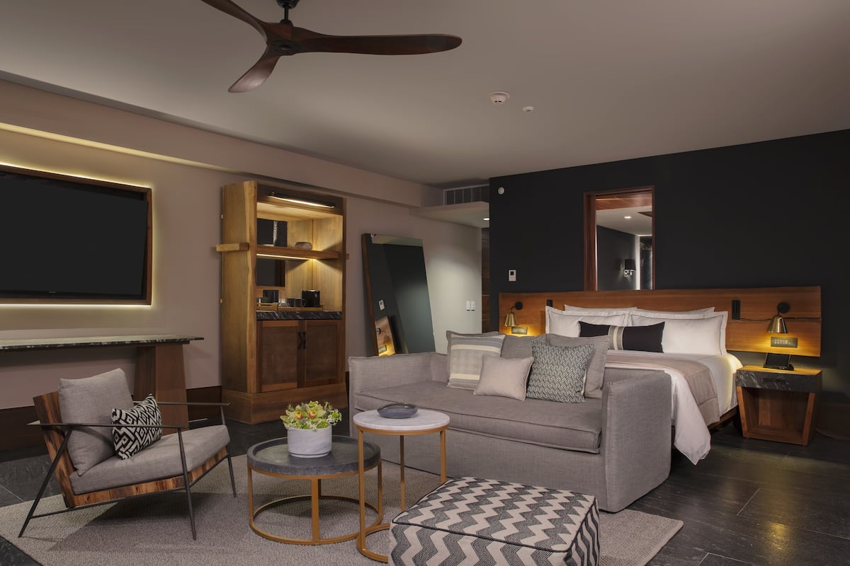 Una habitación de hotel moderna con una cama grande, un sofá, una silla, una mesita y un ventilador de techo.