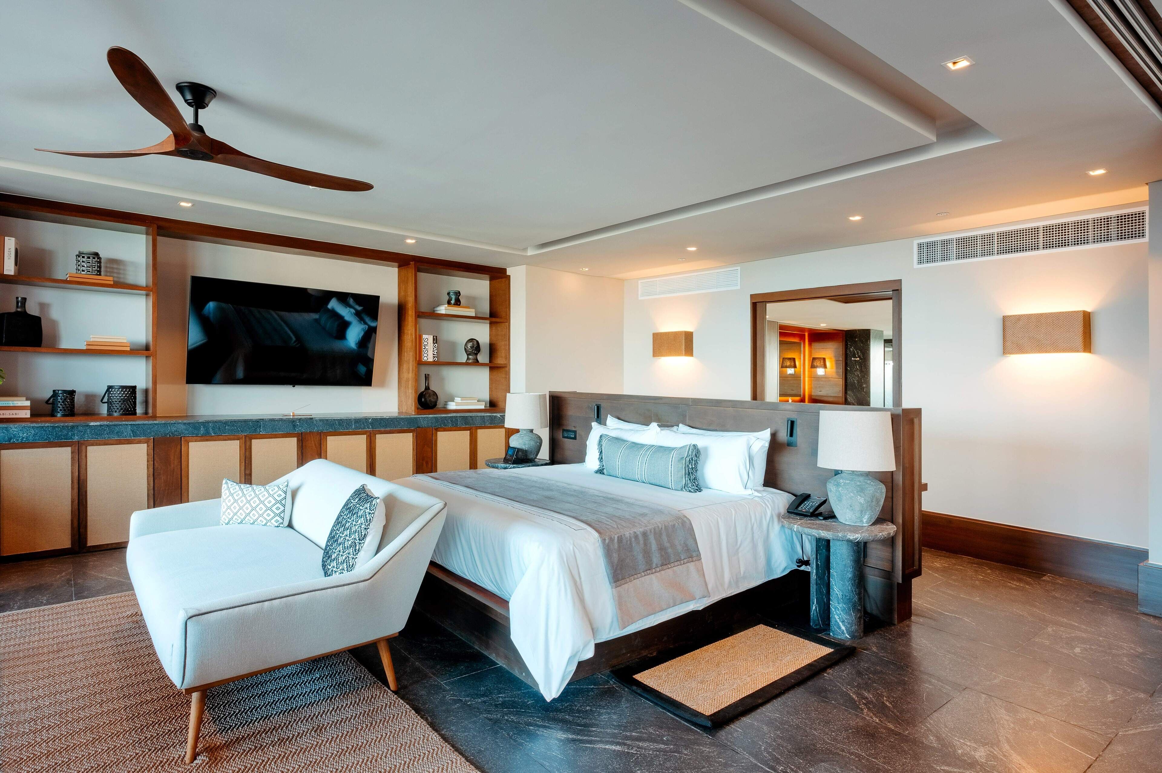 A modern bedroom with a large bed, a TV mounted on the wall, a ceiling fan, and a built-in shelf with various items.