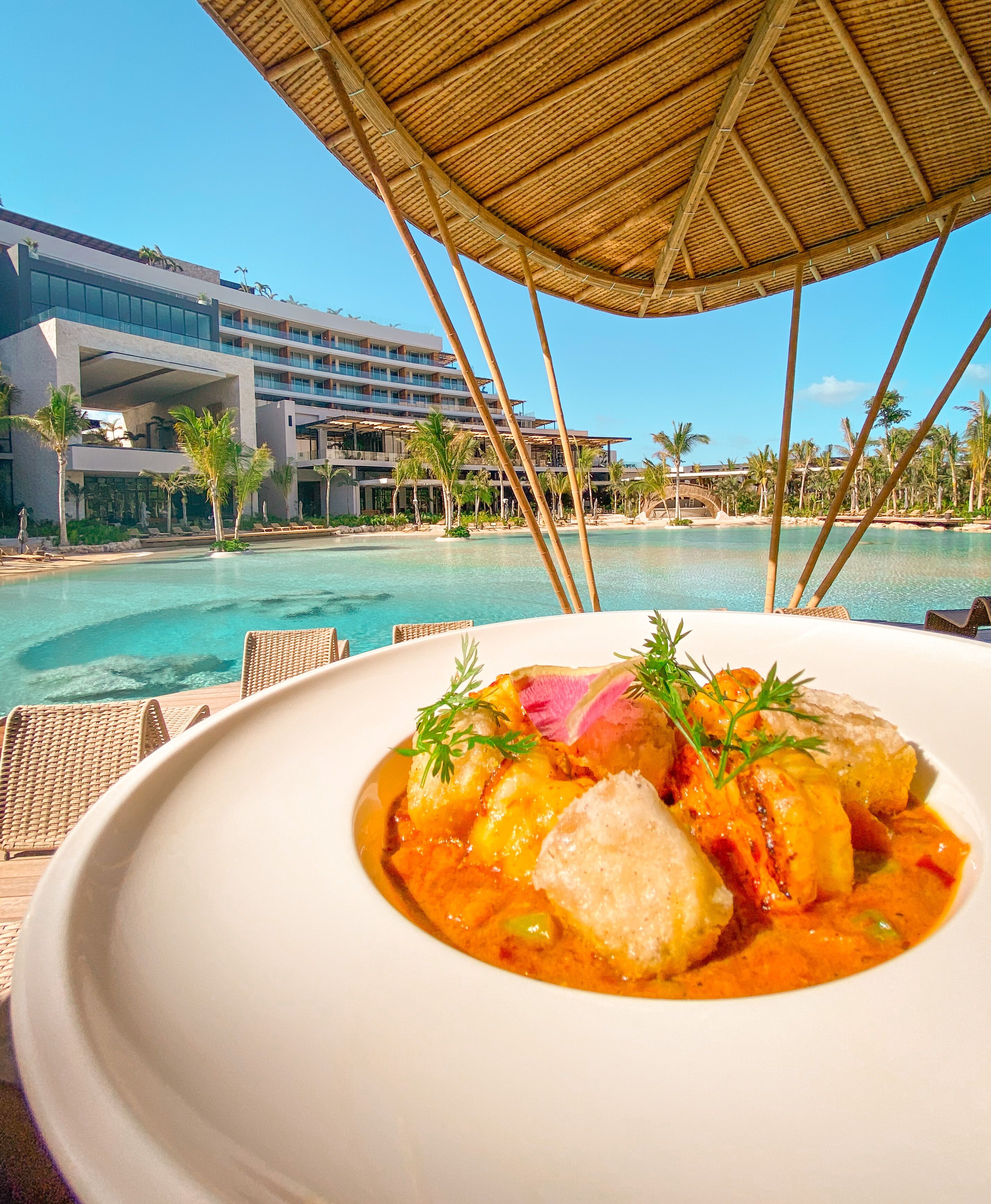 A dining table with a white plate of food, a poolside view, and a resort building in the background.