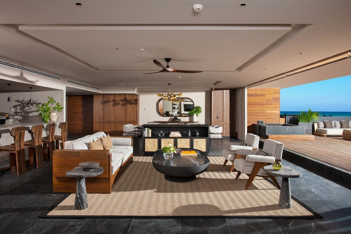 Una sala de estar moderna con sofá, mesa de centro y sillones, un área de cocina con comedor, y un balcón con vista al océano.