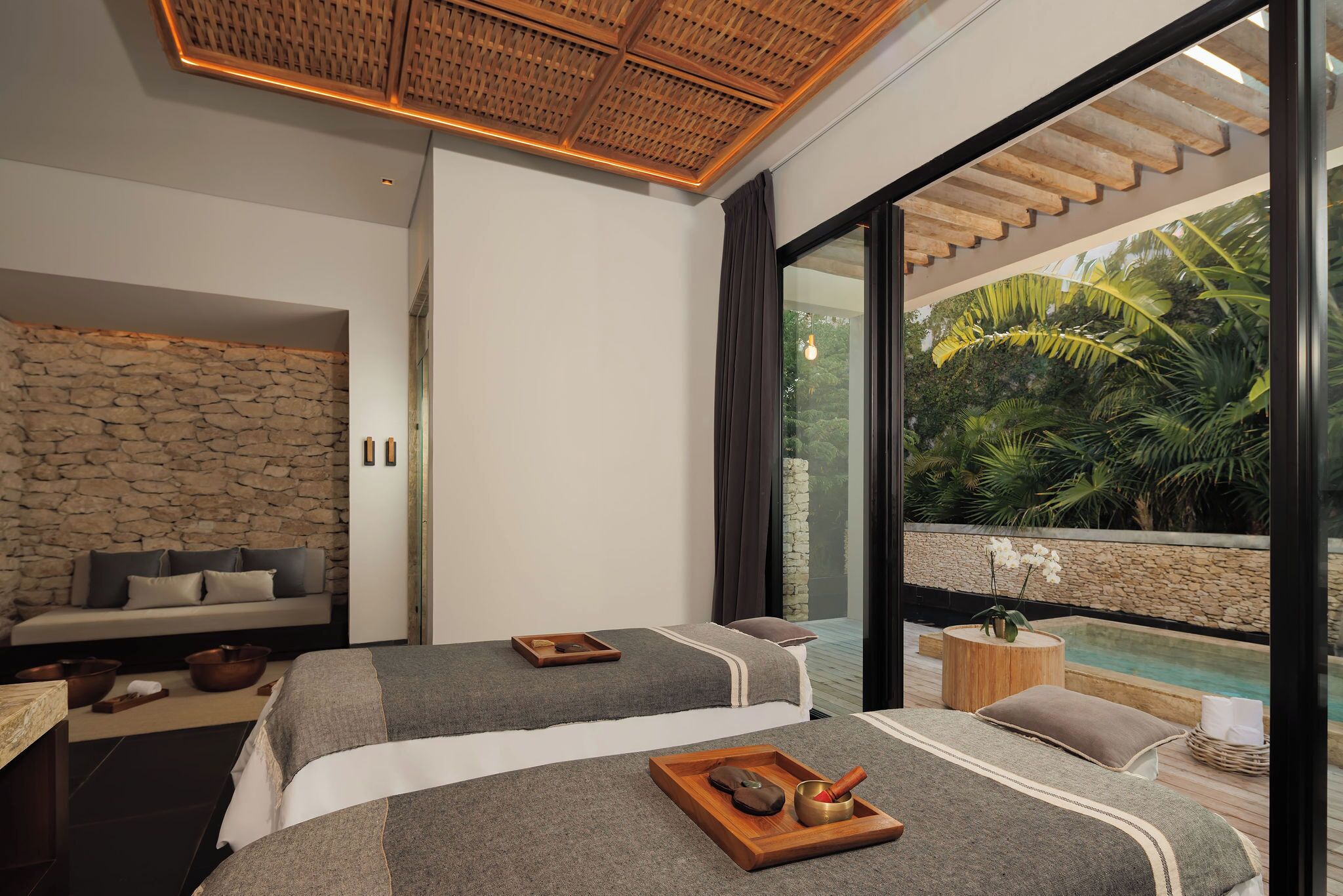 Une chambre moderne avec deux lits, un mur en pierre, un plafond en bois et une vue sur une piscine et la verdure.