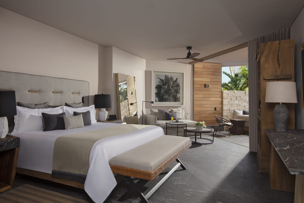 Una habitación de hotel moderna con una cama grande, una zona de estar con una mesa y sillas, un ventanal con vista a palmeras y un ventilador de techo.