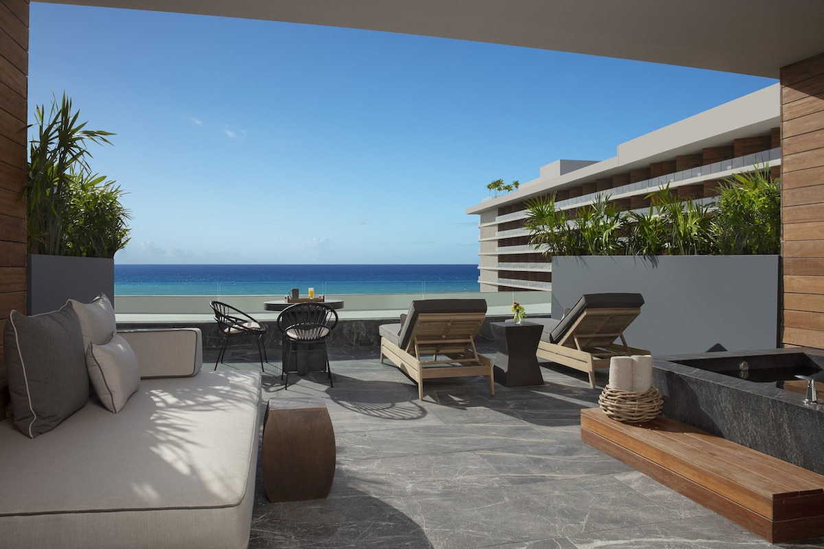 Ein moderner Außenbereich zum Entspannen mit einer Couch, Sesseln, einem Tisch und einem Whirlpool, mit Blick auf einen Strand.