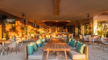 A spacious outdoor seating area with a long wooden table and multiple chairs, surrounded by cushioned sofas and pillows.