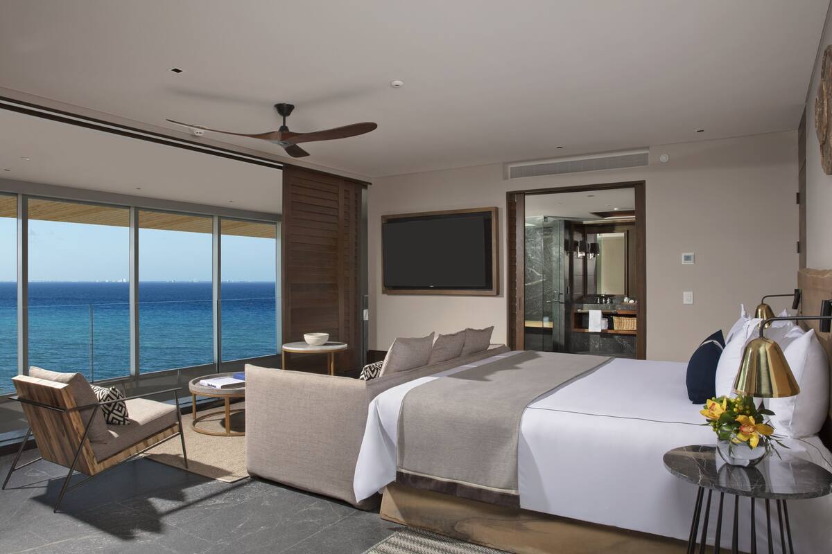 Une chambre d’hôtel moderne dotée d’un grand lit, d’un coin salon avec vue sur l’océan, d’un ventilateur de plafond et d’une salle de bain avec un grand miroir.