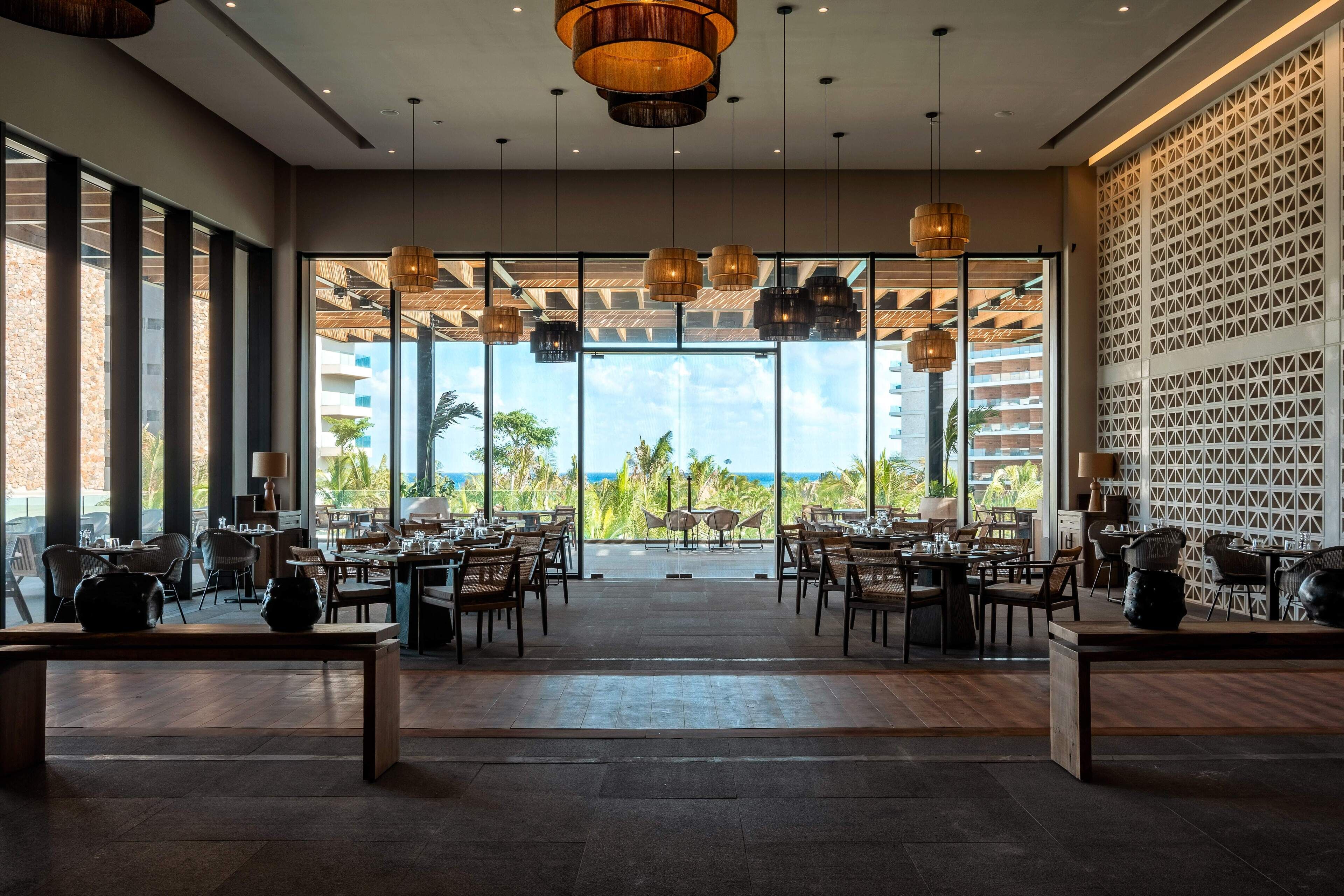 A spacious dining area with multiple tables and chairs, large windows offering a view of the ocean, and a patterned wall on the right.