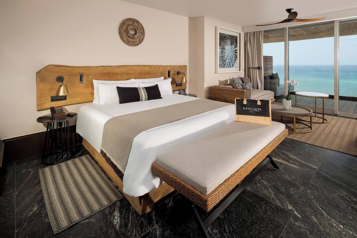 Une chambre d’hôtel avec un grand lit, une table de chevet avec une lampe, une commode, une petite table avec une chaise, et une vue sur l’océan grâce à de grandes fenêtres.