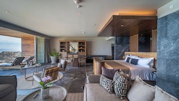 A modern hotel room with a large bed, a seating area with a sofa and chairs, a dining table, and a view of the ocean.