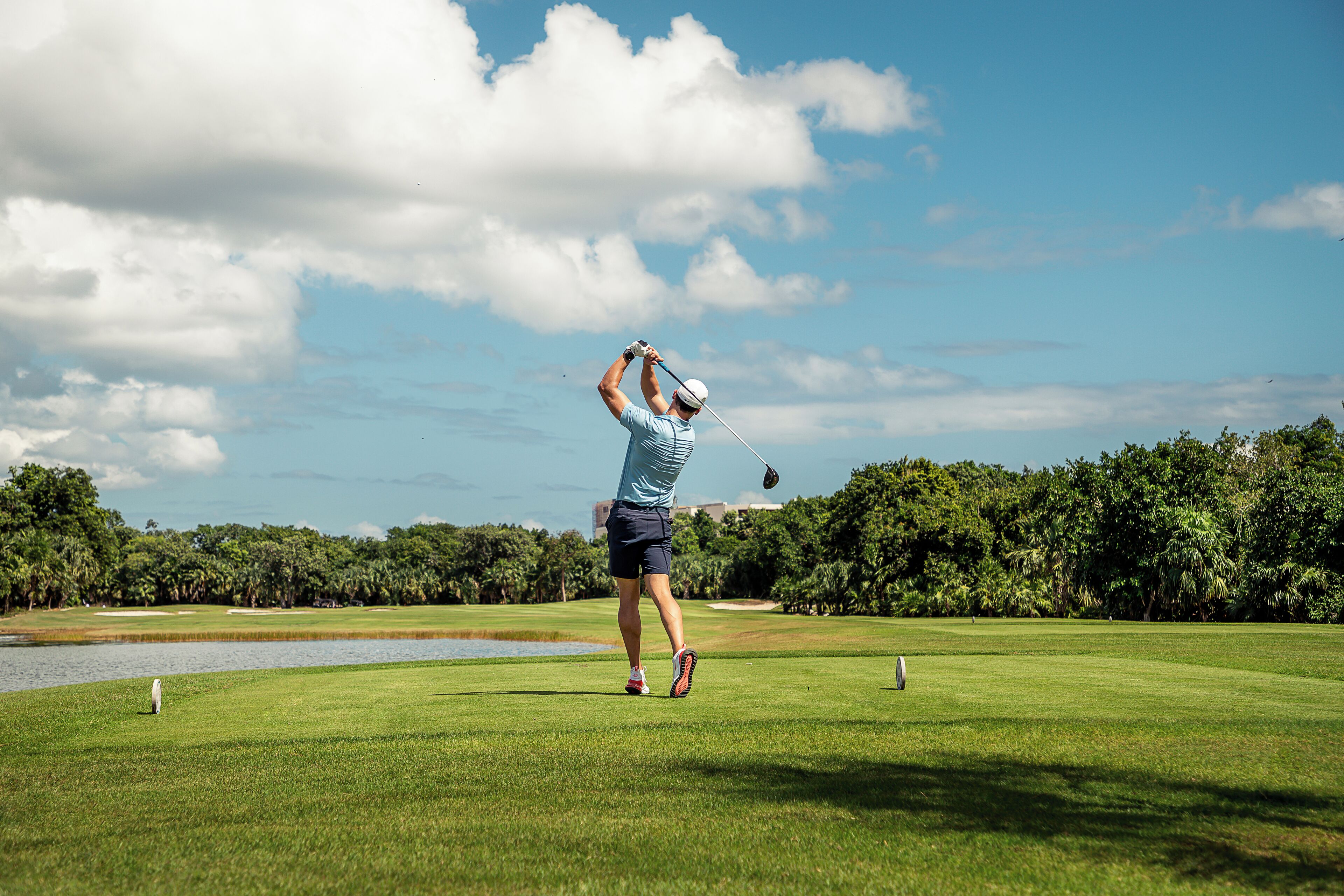 A golfer in mid-swing on a golf course with a clear sky and lush greenery in the background.