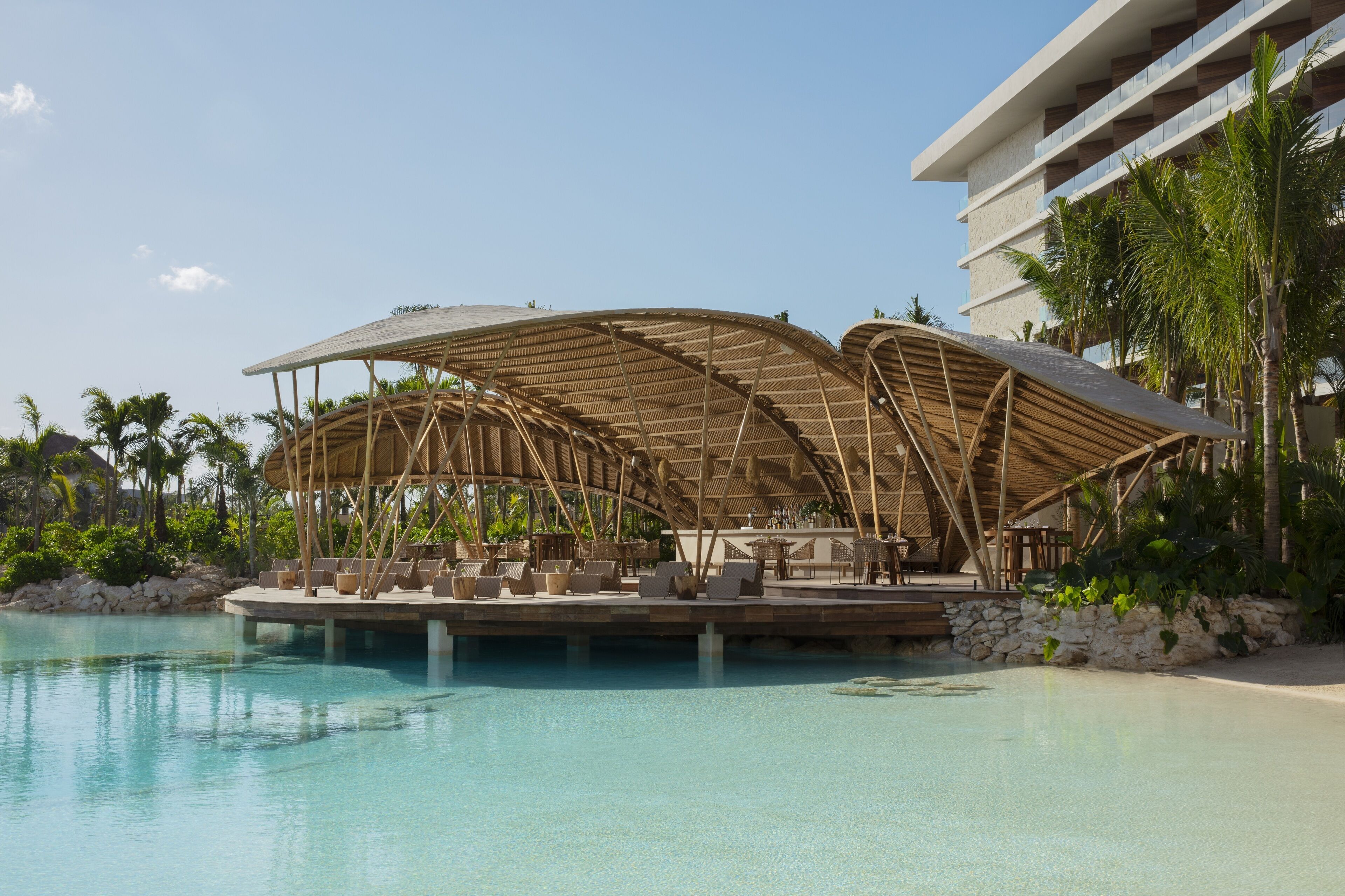 A resort pool area with bamboo canopies, lounge chairs, and a building in the background.
