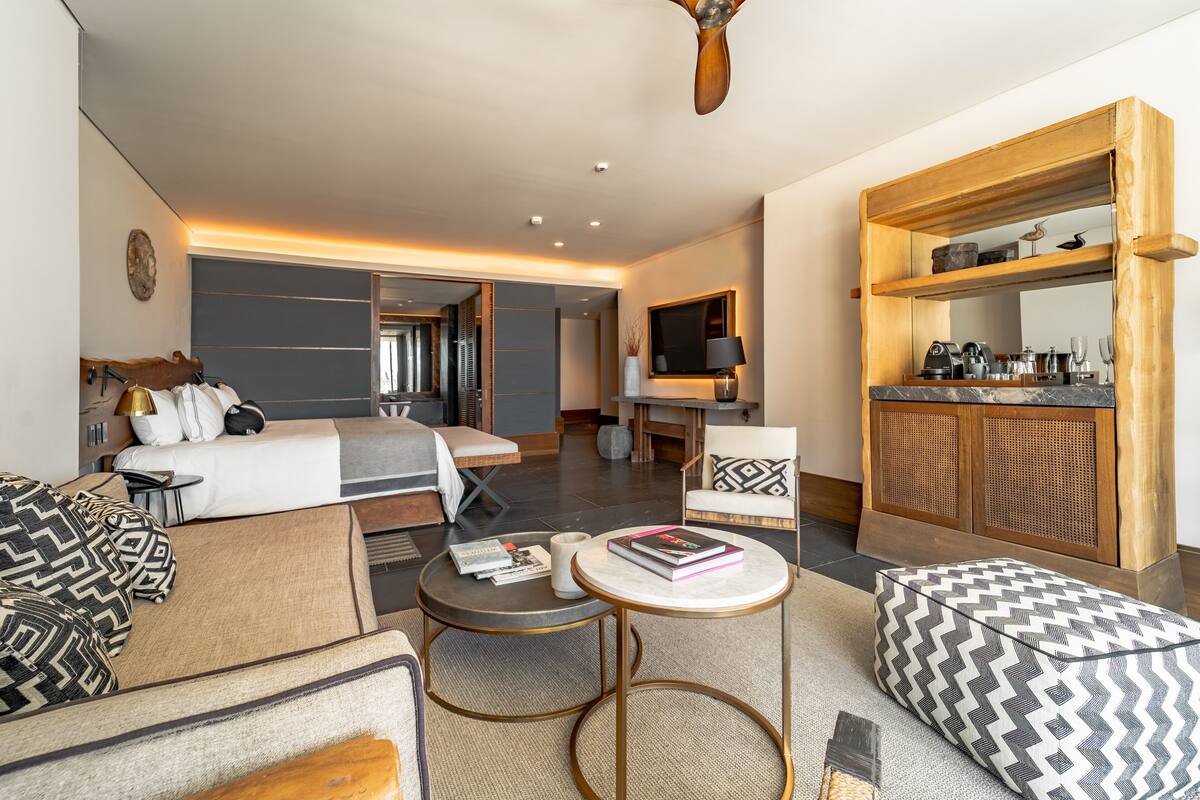 Une chambre d’hôtel moderne comprenant un canapé, une petite table ronde, un lit avec une tête de lit, une télévision, une armoire en bois et un grand miroir.