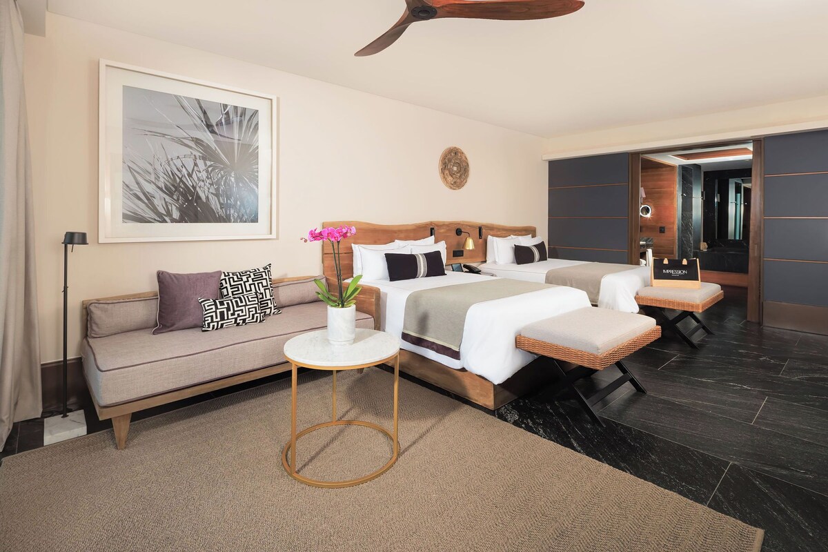 Una habitación de hotel moderna con una cama grande, un sofá, una pequeña mesa redonda con una planta y un ventilador de techo.