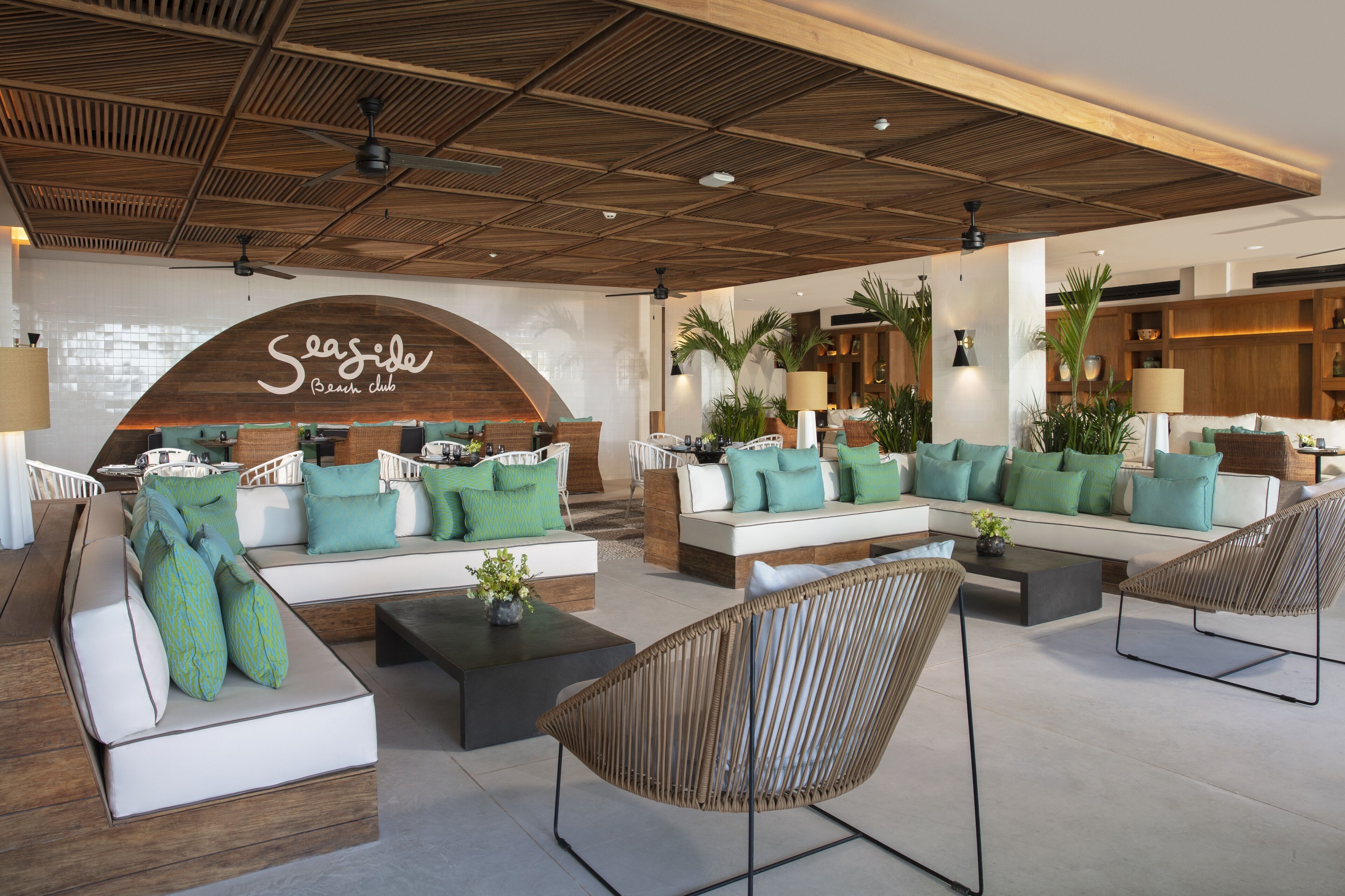 Amplia zona de estar con techo de madera, varias opciones de asientos que incluyen sofás y sillones, y un llamativo letrero que dice 'Seaside Beach club'.