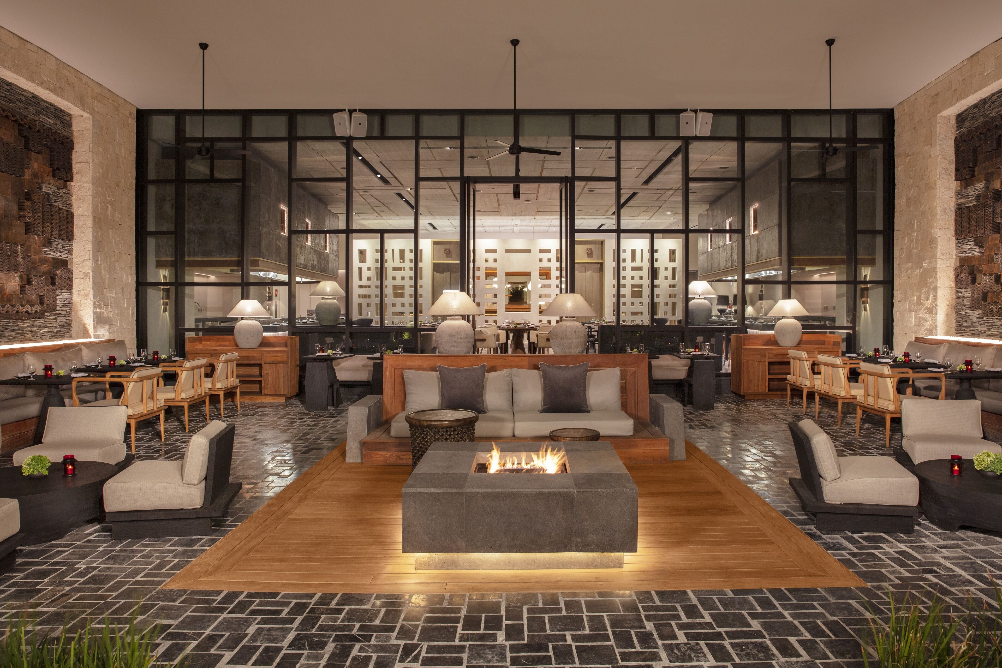 A modern lounge area with a central fire feature, seating arrangements, and a view of the kitchen through large windows.