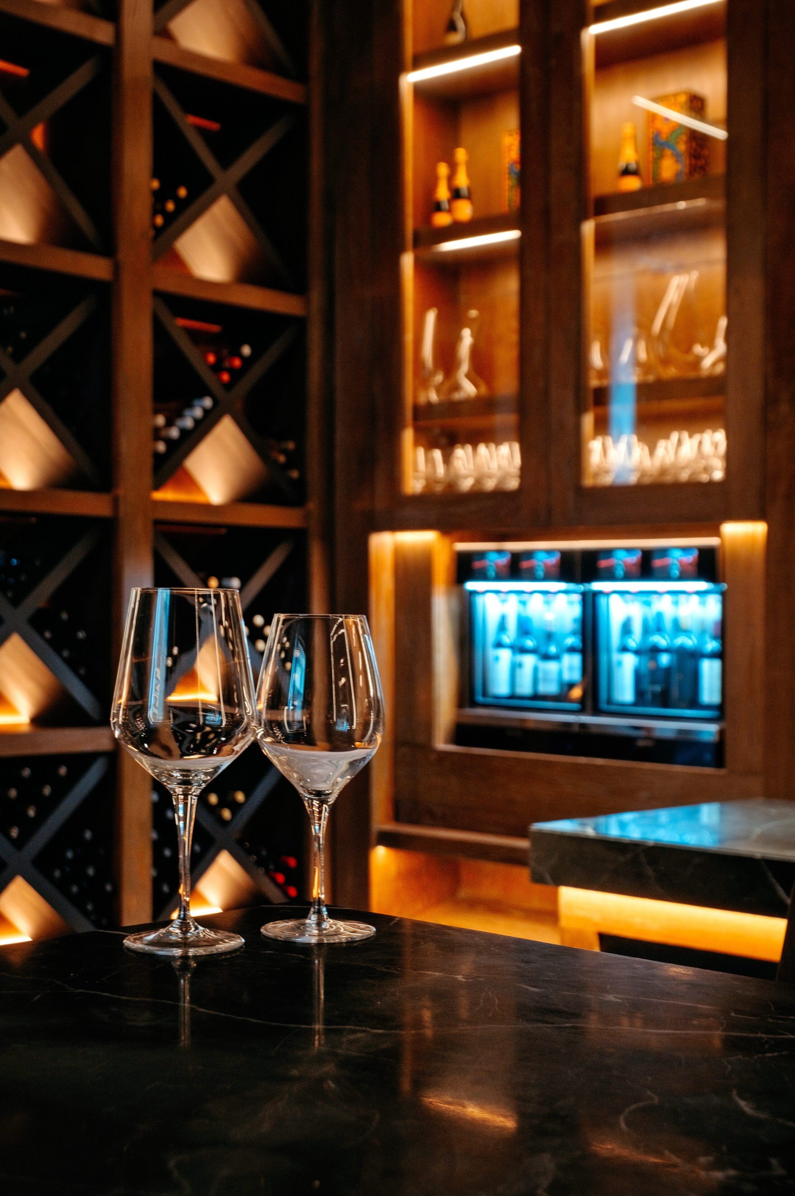 Dos copas de vino vacías sobre una barra, con una alacena para vinos y estantes al fondo.
