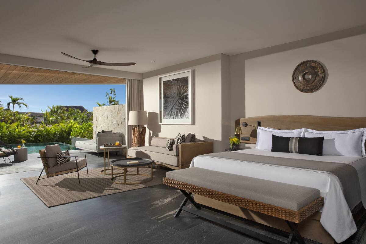 Une chambre d’hôtel moderne dotée d’un grand lit, d’un canapé, d’un fauteuil, d’une petite table et offrant une vue sur une piscine et un espace verdoyant.