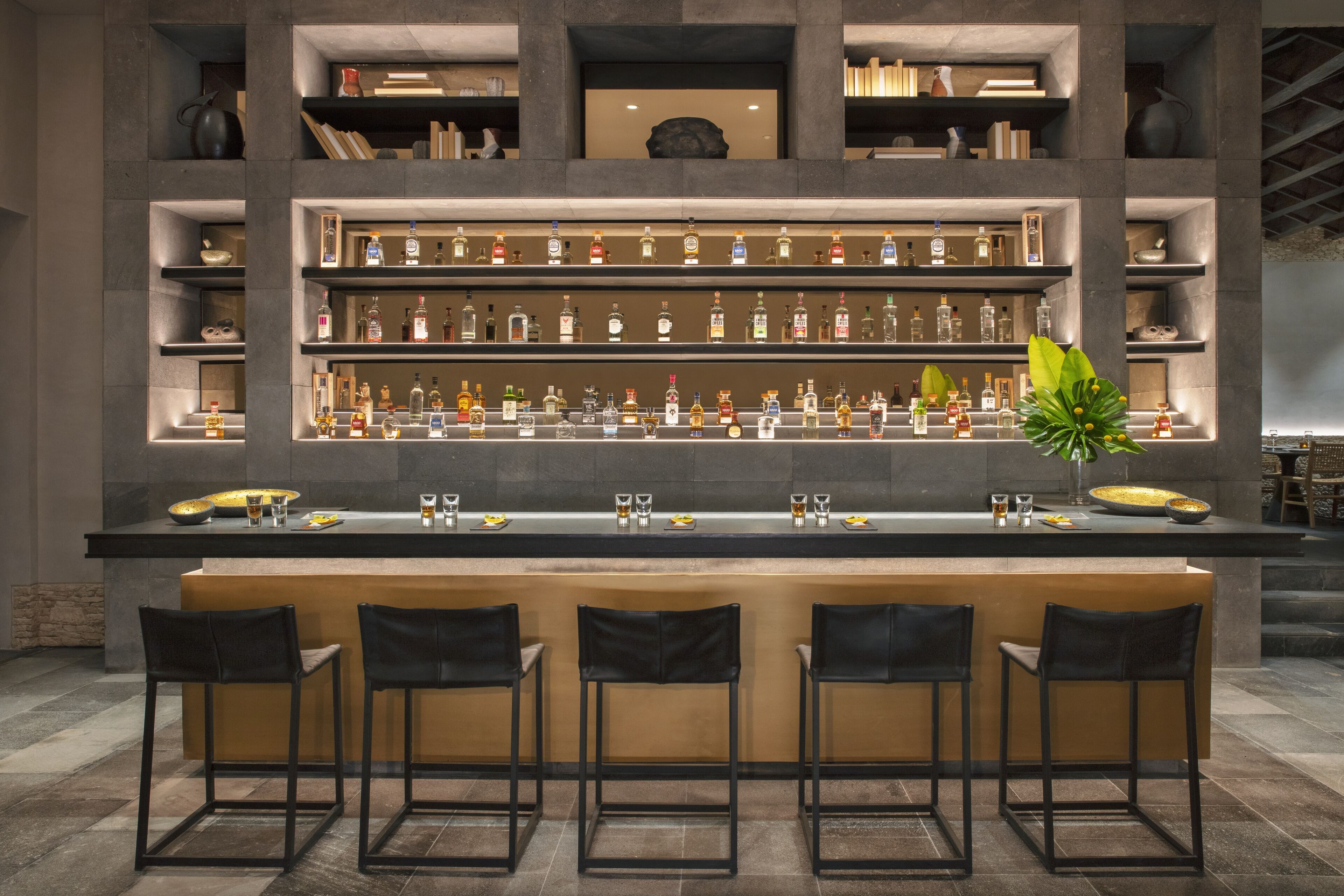 Una zona de bar con varias estanterías llenas de botellas, una barra central y cuatro taburetes de bar.