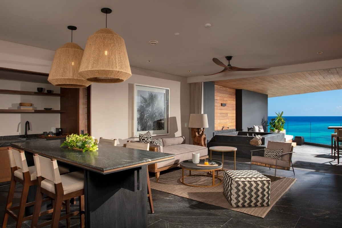 Una sala de estar moderna con una isla grande, taburetes de bar, un sofá y vista al océano.