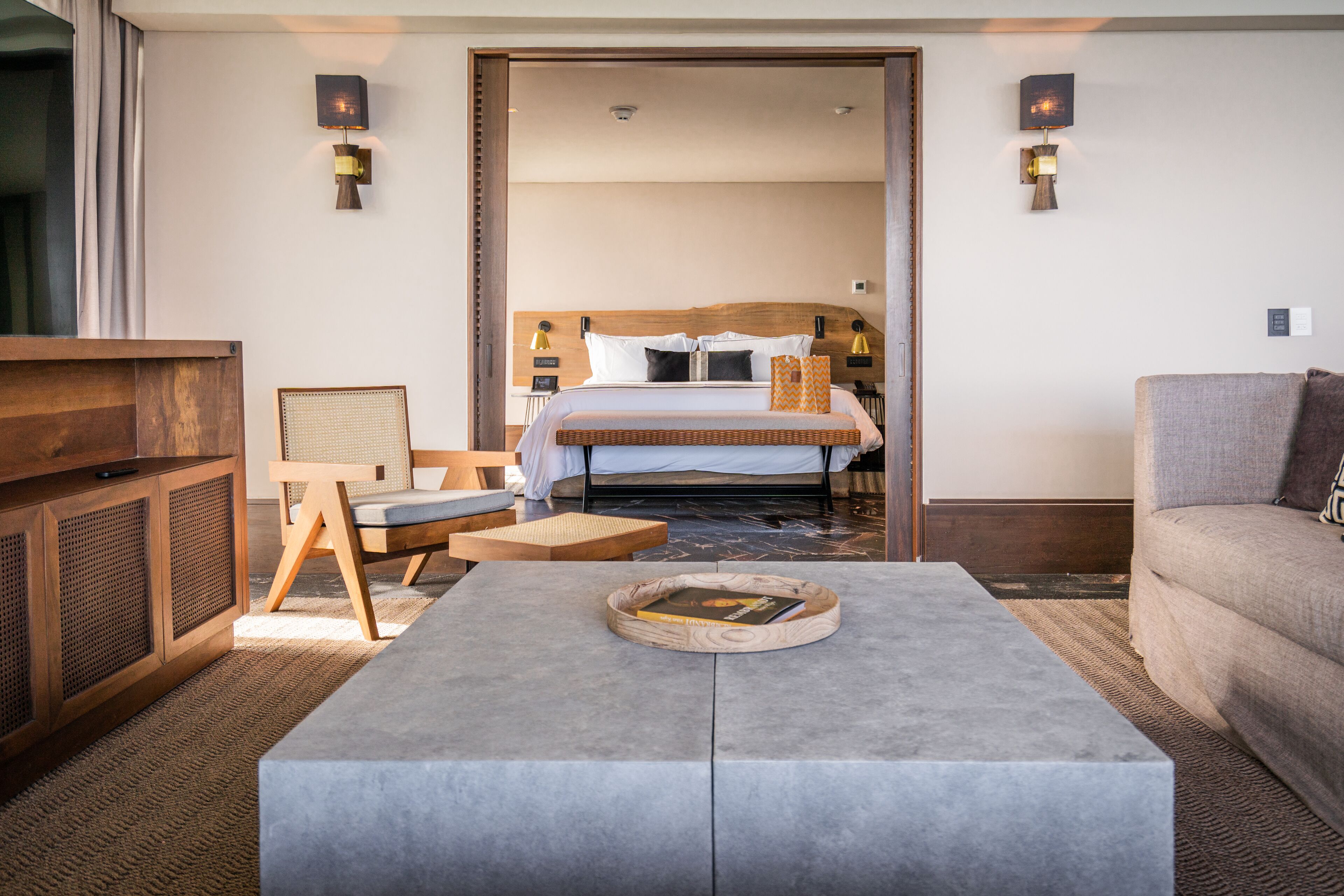 A hotel room with a bed, a wooden nightstand, a grey sofa, a coffee table, and a TV.