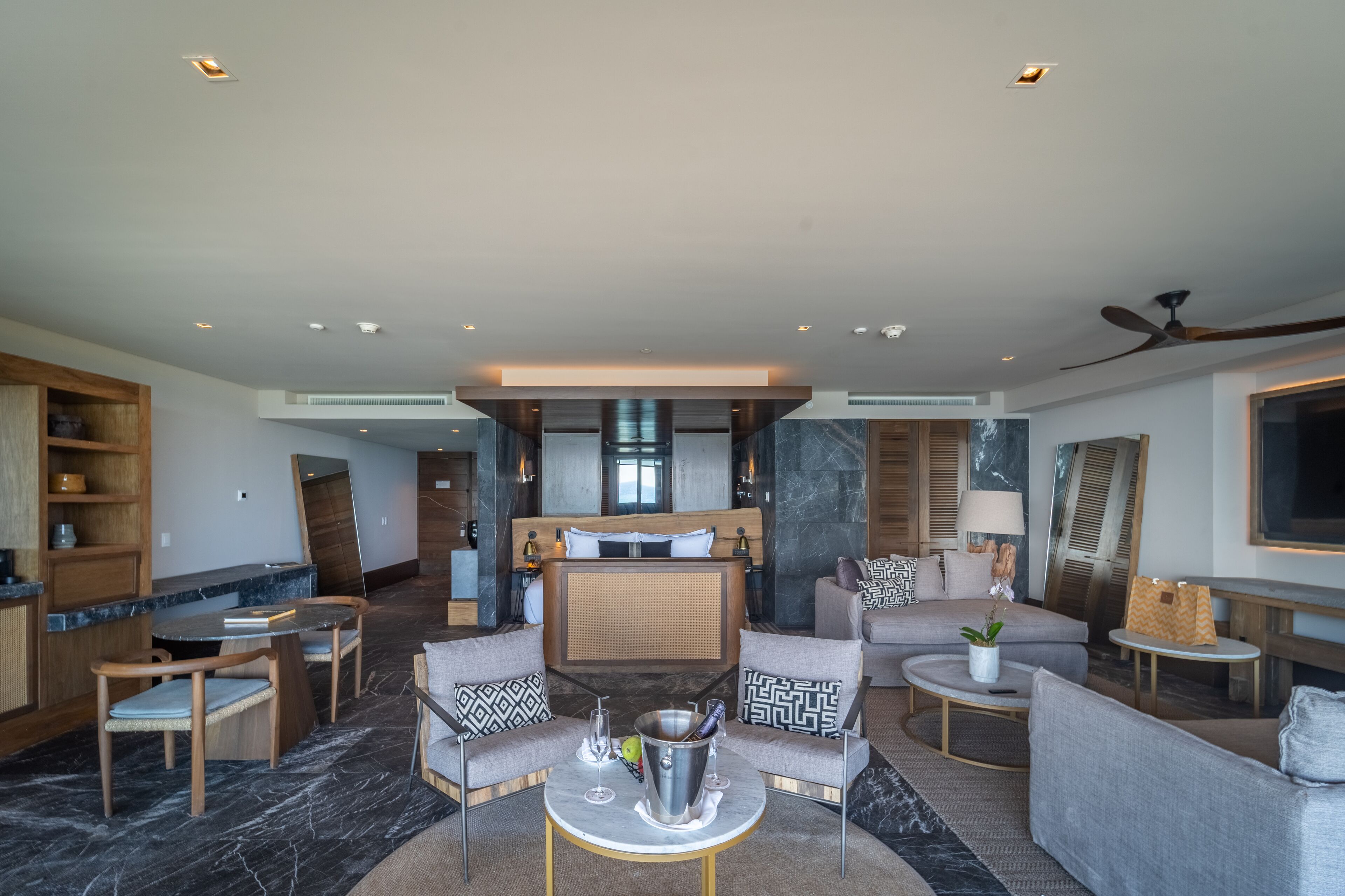 A modern hotel room with a large bed, a sitting area with sofas and a coffee table, a kitchen area, and a large TV on the wall.