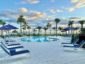 A pool area with lounge chairs and umbrellas, surrounded by palm trees and a view of the ocean.