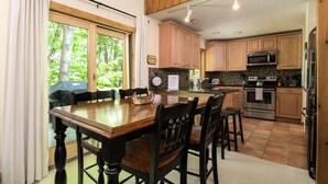 A kitchen with wooden cabinets, a dining table with chairs, a clock, and a view of trees through the window. - Mountain condo by Okemo w/ hot tub (Ludlow)