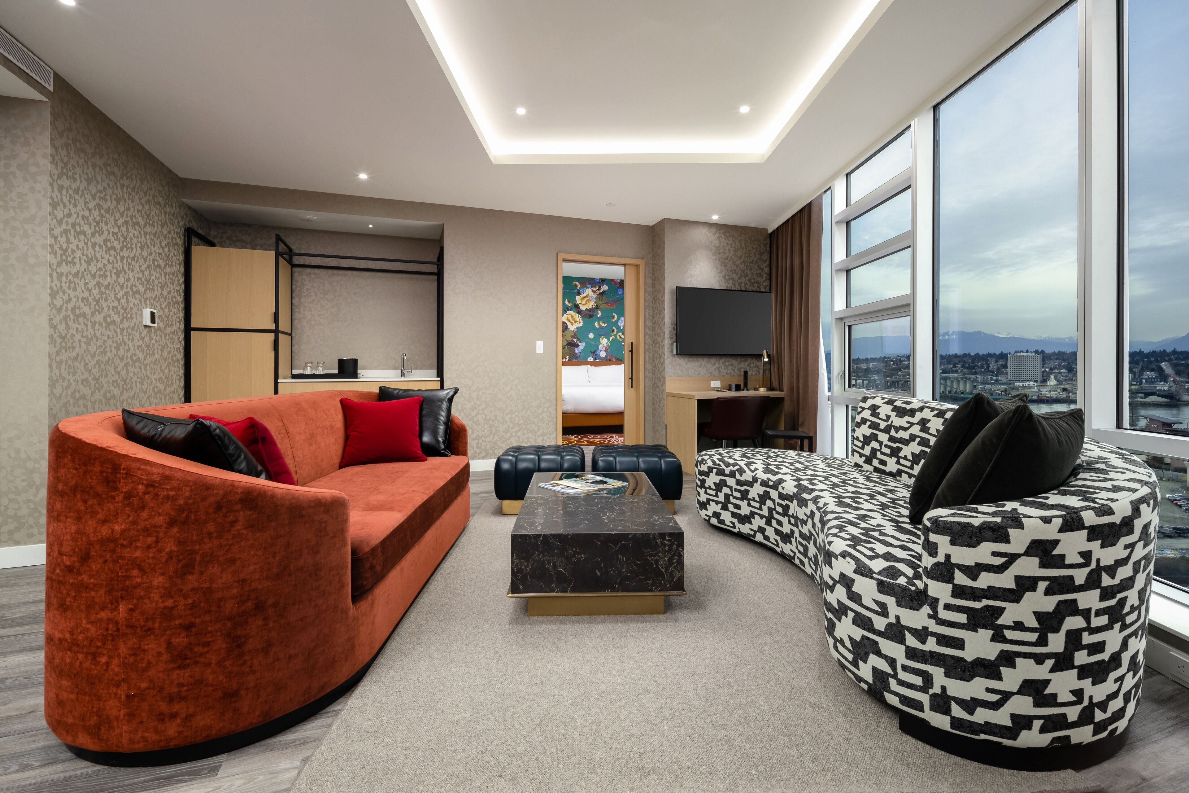 A modern living room with a curved orange sofa, a patterned black and white sofa, and a black coffee table. There is a kitchen area with a wooden cabinet and a flat-screen TV mounted on the wall.