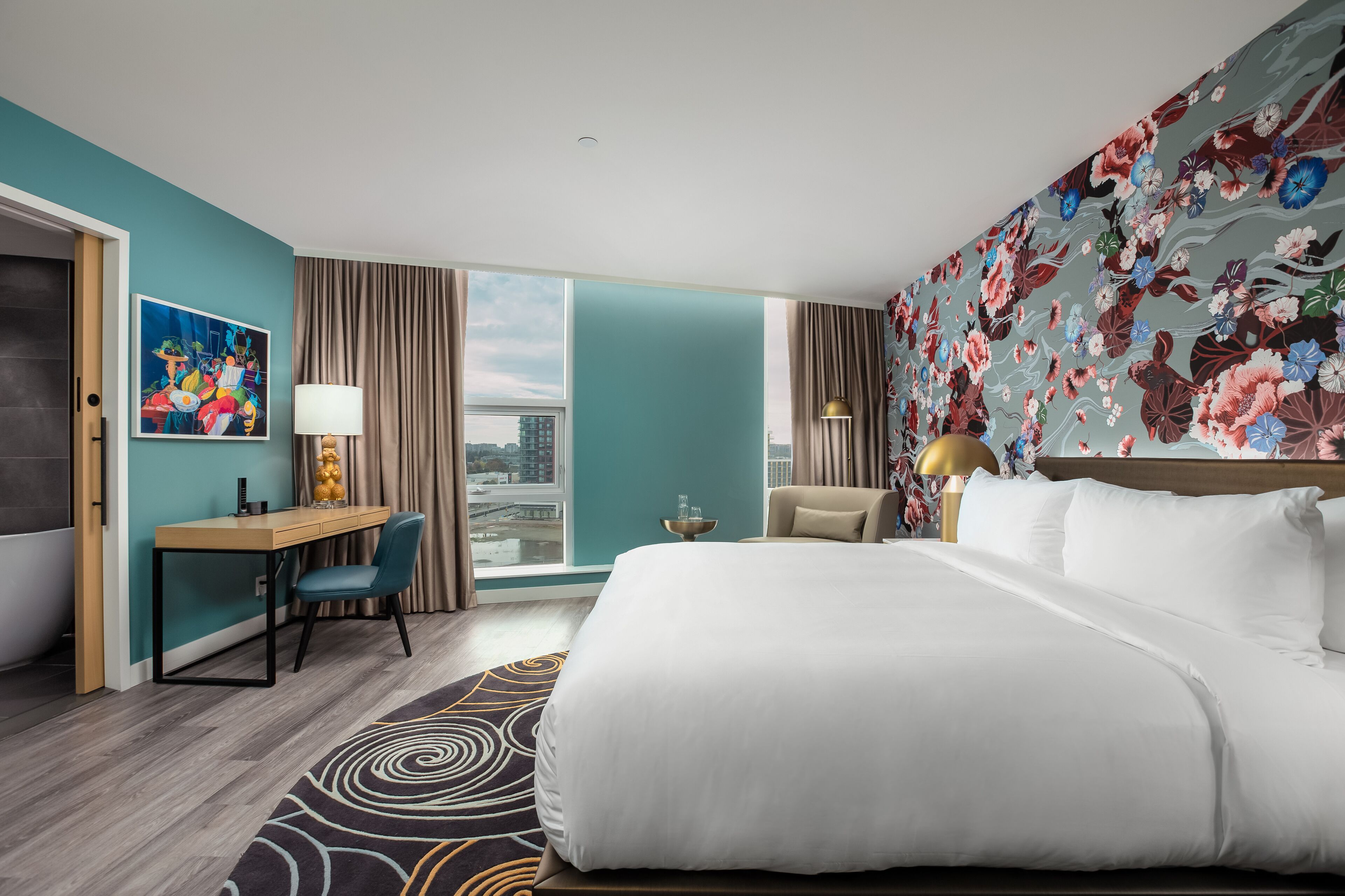 Quarto de hotel com uma cama grande, uma escrivaninha com cadeira, vista para a cidade e parede decorada com flores coloridas.