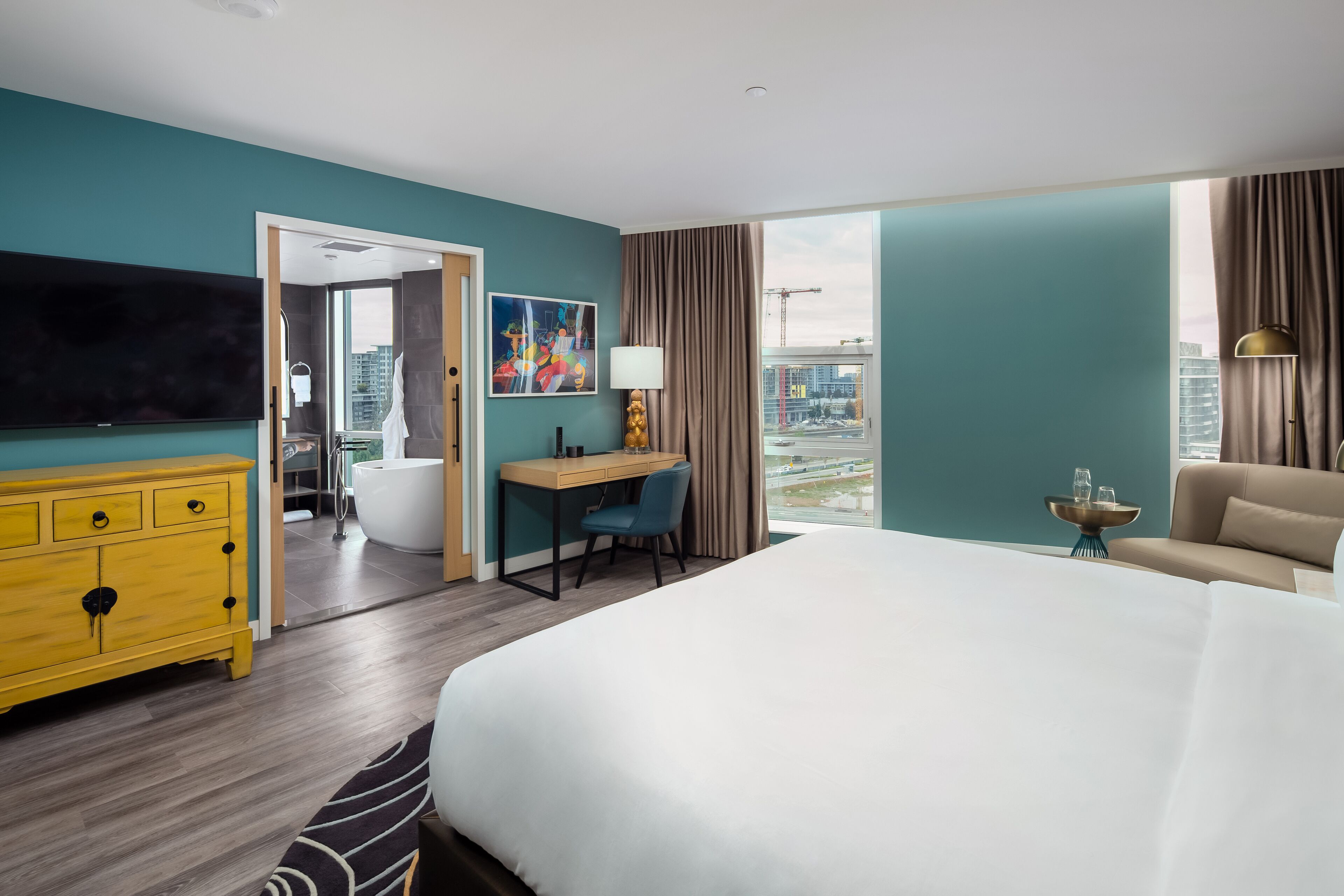 A hotel room with a large bed, a desk, a chair, a TV, a bathroom, and a view of the city.
