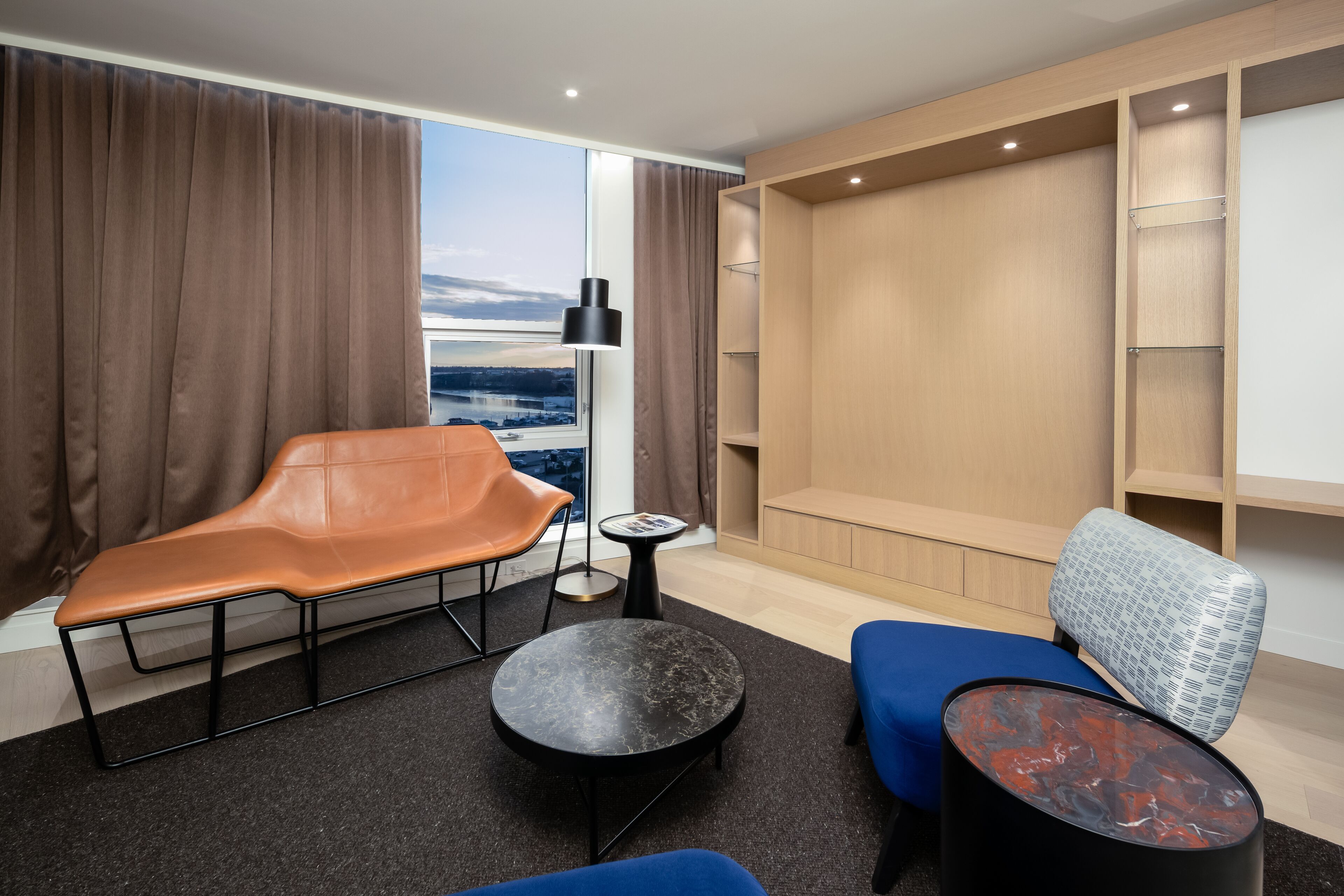 Moderni hotellihuone, jossa on suuri ikkuna, oleskelualue sohvineen ja tuoleineen, pieni pyöreä pöytä sekä seinään upotetut hyllyt.