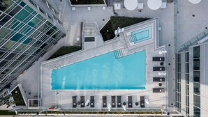 An aerial view of a modern building with a large swimming pool, outdoor seating areas, and a rooftop garden.