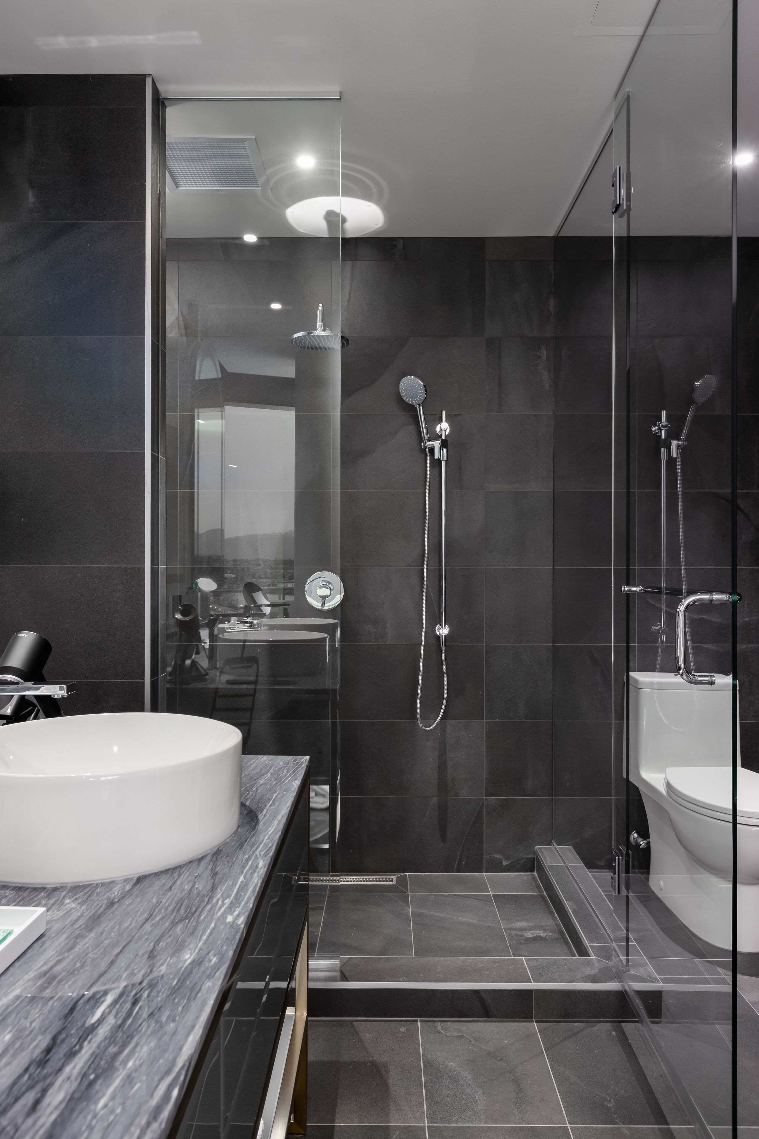 A modern bathroom with a large white sink, a glass shower enclosure, and dark tiled walls.