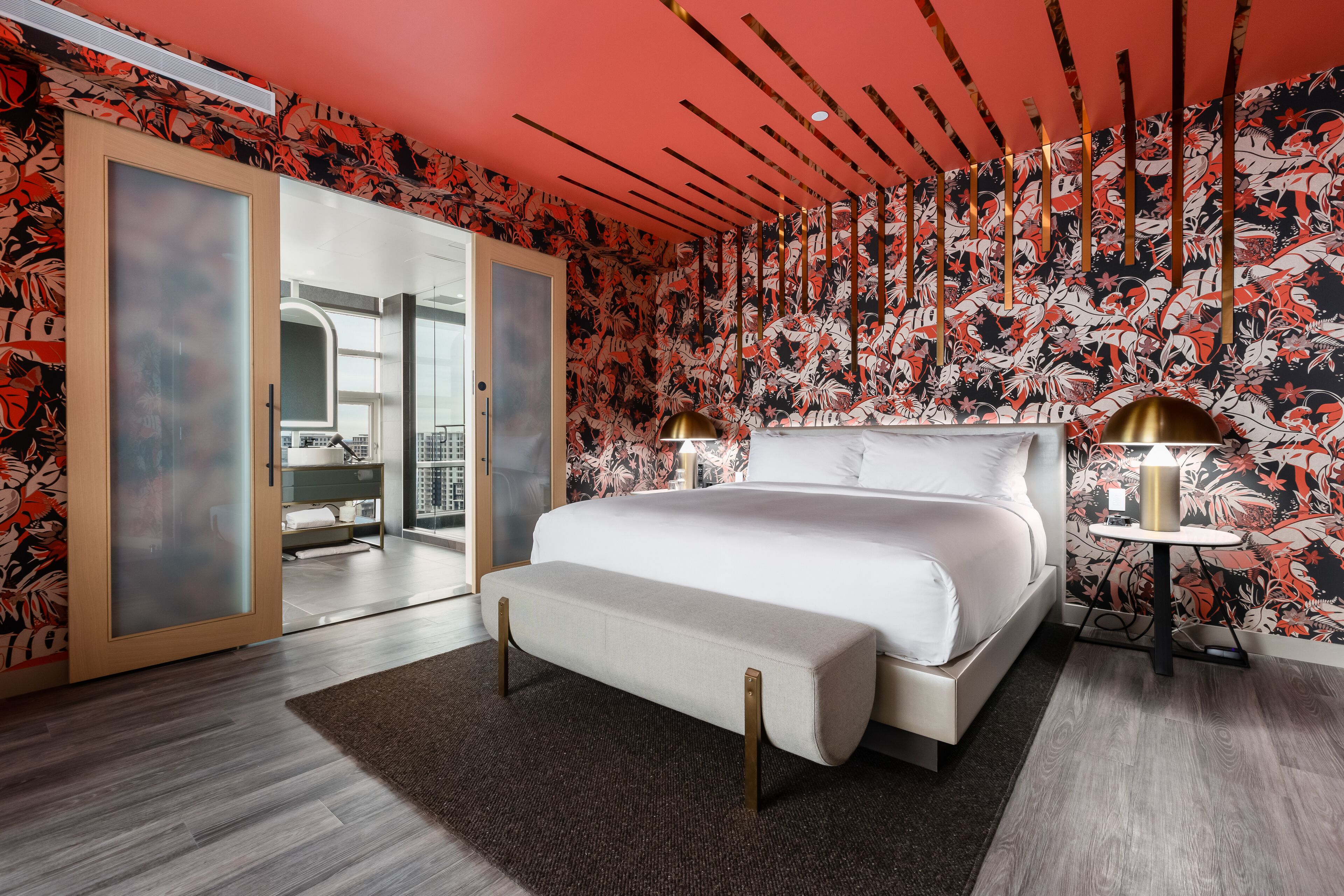 Un dormitorio moderno con una cama grande, una banca, una mesita de noche con lámparas y vistas a un paisaje urbano a través de una puerta corredera.