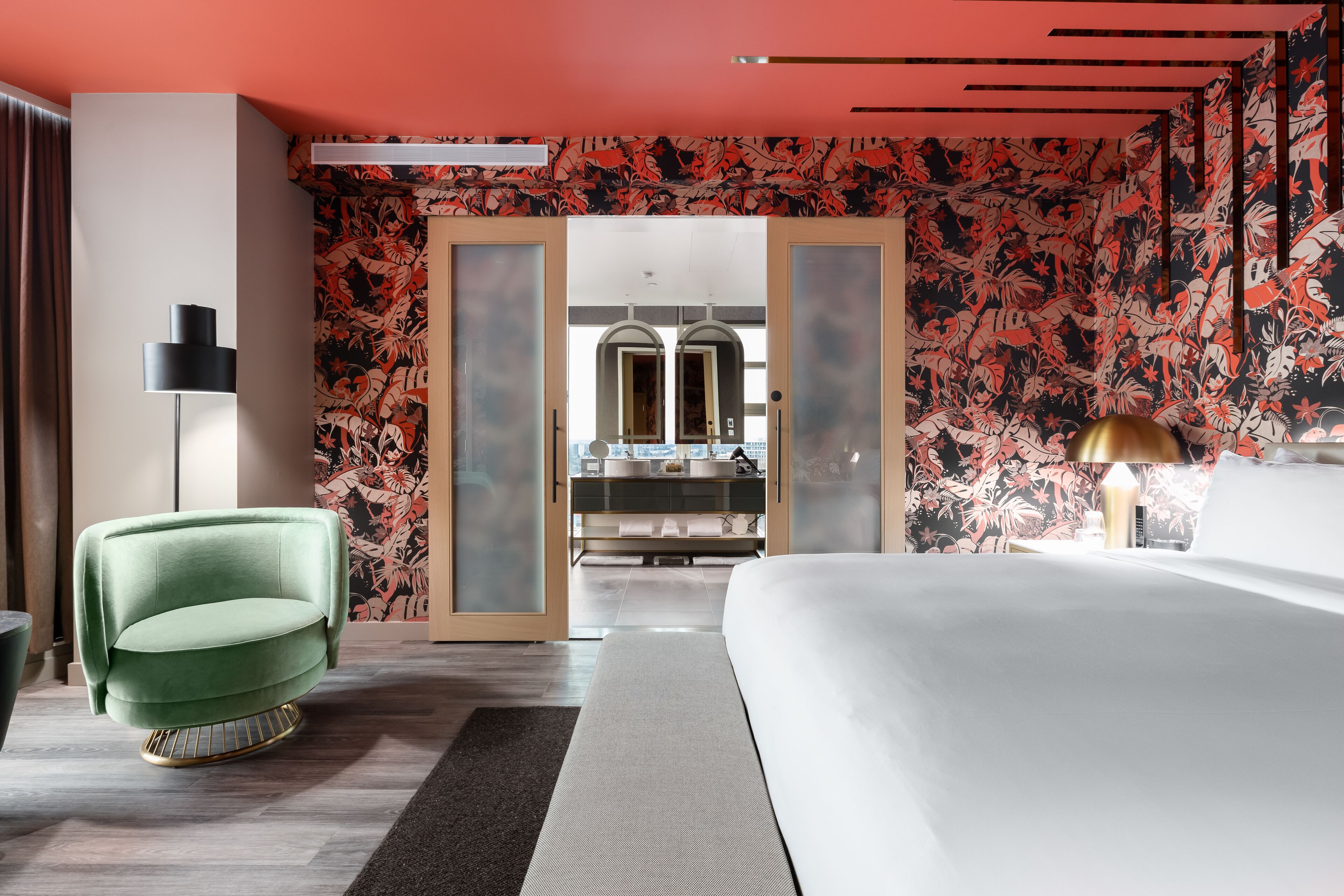 Une chambre d’hôtel moderne avec un grand lit, un fauteuil vert, une lampe sur pied et une salle de bain visible à travers une porte en verre.