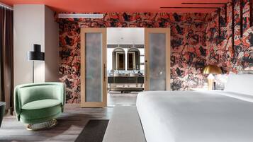 Une chambre d’hôtel moderne avec un grand lit, un fauteuil vert, une lampe sur pied et une salle de bain visible à travers une porte en verre.
