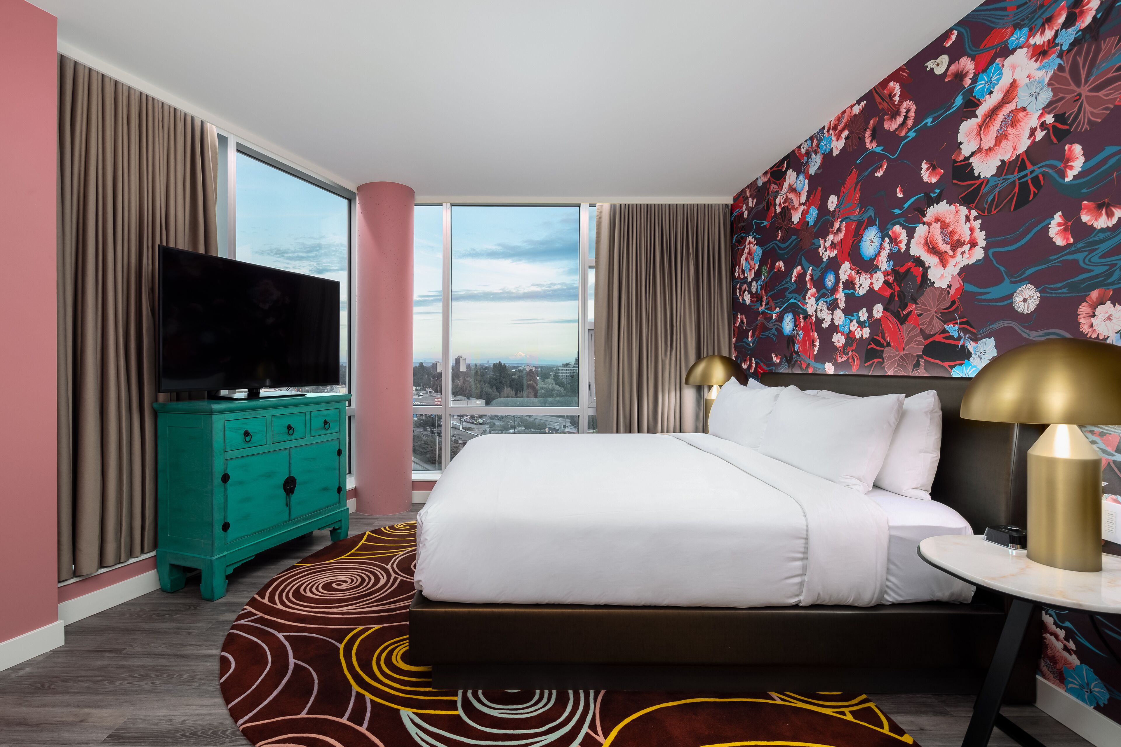 A hotel room with a large bed, a television on a teal dresser, a round table with a lamp, and a floral wallpaper.