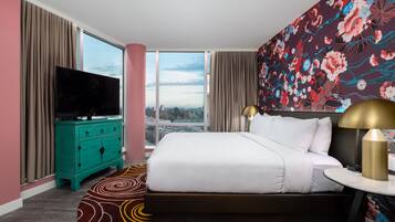 A hotel room with a large bed, a television on a teal dresser, a round table with a lamp, and a floral wallpaper.