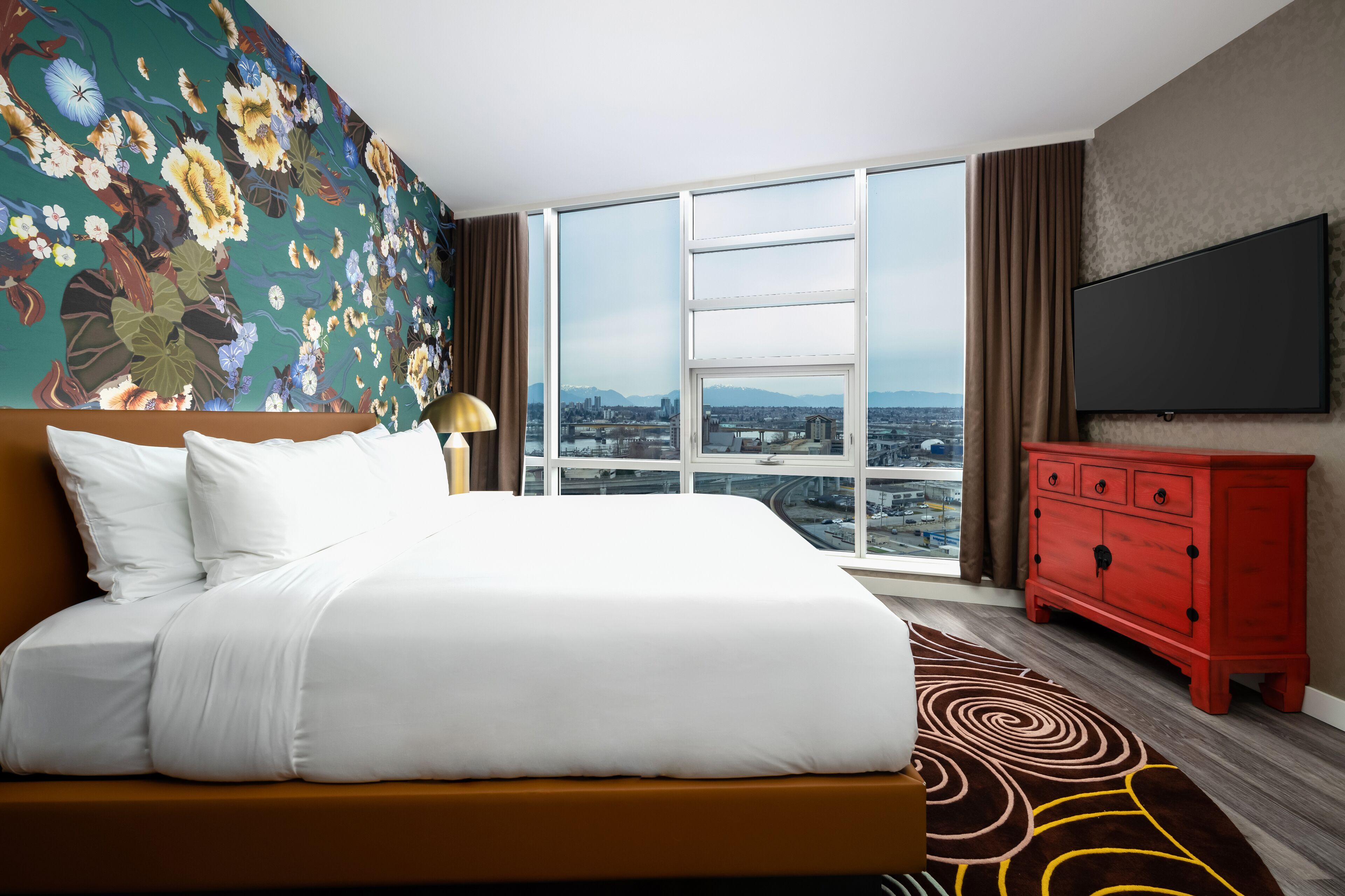 A hotel room with a large bed, a red dresser, a flat-screen TV, and a city view through large windows.