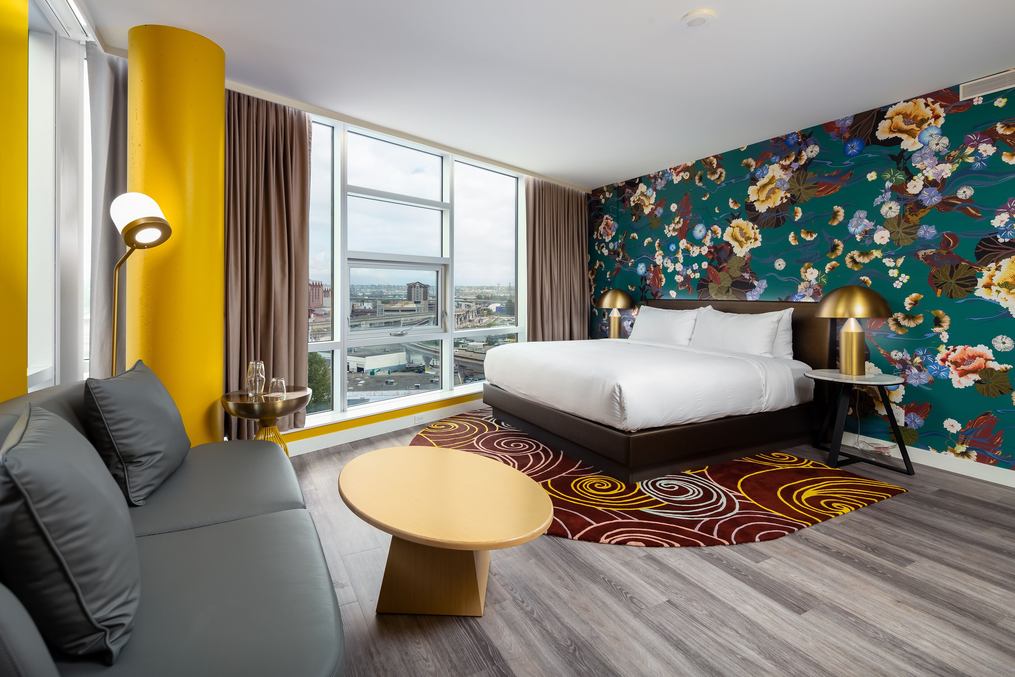 A modern hotel room with a large bed, a sofa, a round table, and a view of the city through large windows.