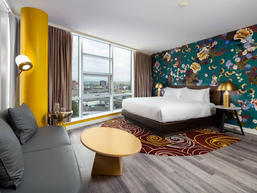 A modern hotel room with a large bed, a sofa, a round table, and a view of the city through large windows.