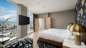 A modern hotel room with a large bed, a flat-screen TV, a desk with a lamp, and a view of the city through large windows.