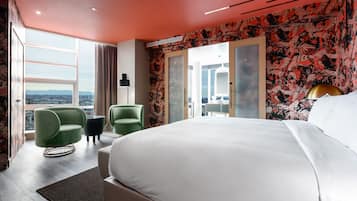 A modern hotel room with a large bed, a green armchair, a small table, and a view of the city through large windows.