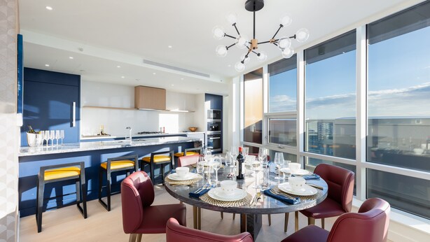A modern kitchen with a central island, large windows, and a dining area with round tables and red chairs.
