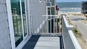 Una terraza de madera con barandal blanco, puerta de cristal y vista a la playa y al océano.
