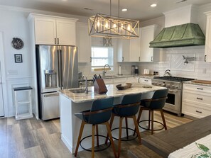 A modern kitchen with a central island, stainless steel appliances, and bar stools. - Stone's Throw - Newly Constructed 4 Bedroom w/heated pool - 1 Block to the Ocean (Ocean Isle Beach)