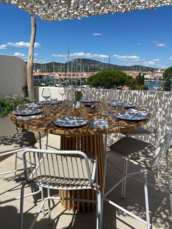 Une table ronde en bois, dressée pour six personnes, entourée de chaises en métal blanc, et offrant une vue sur une marina avec des bateaux et des bâtiments en arrière-plan.
