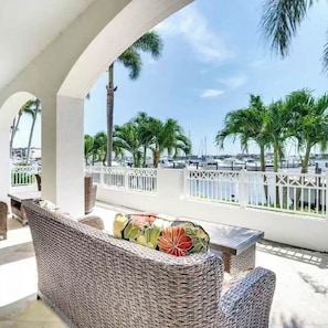 A covered outdoor seating area with wicker furniture and a view of a marina with boats and palm trees.