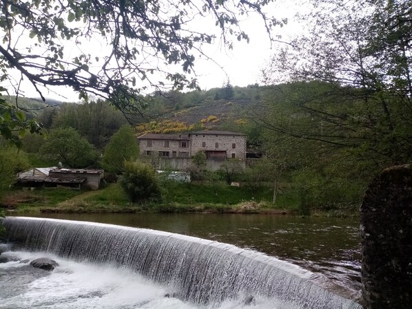 Un bâtiment rustique en pierre, doté d’une architecture traditionnelle, entouré d’une végétation luxuriante et bordé par une rivière aux eaux vives.