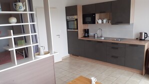 A modern kitchen with grey cabinets, a stainless steel sink, and a microwave. There is a shelf with decorative items and a kettle on the countertop. - Studio Vue mer RDC à 100 m de la Plage (Larmor-Plage)